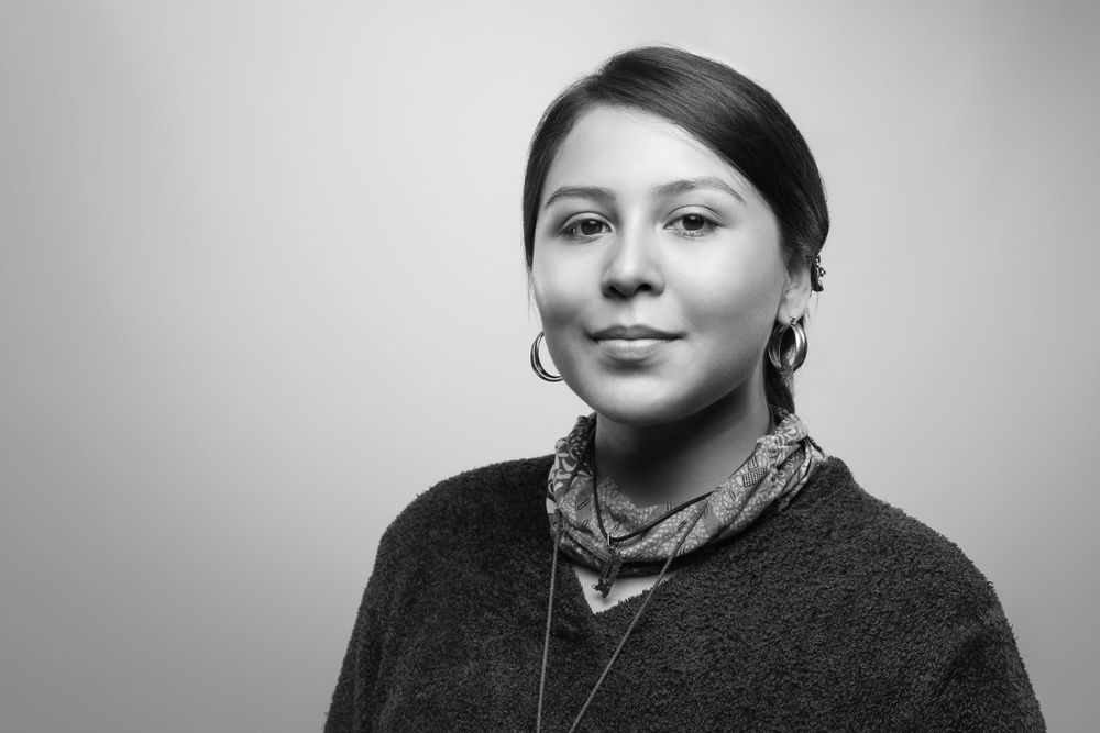 A black and white studio portrait of someone with dark hair wearing a charcoal sweater and patterned scarf, looking at camera.