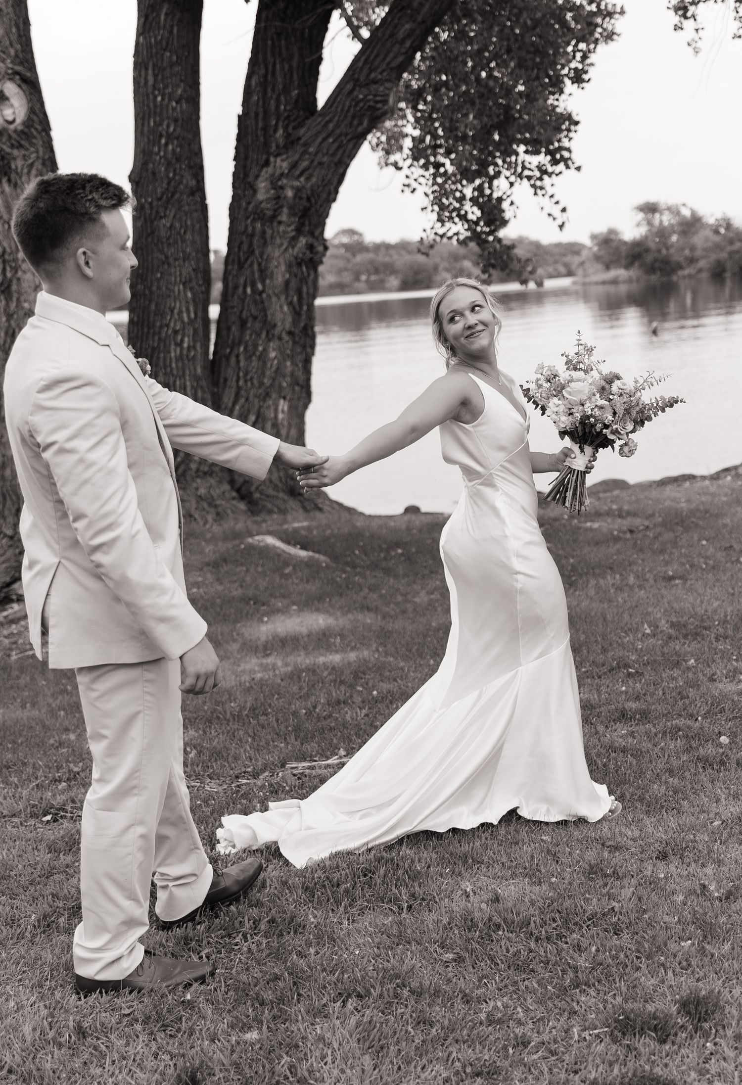 Playful wedding couple dancing by a lake under large trees.