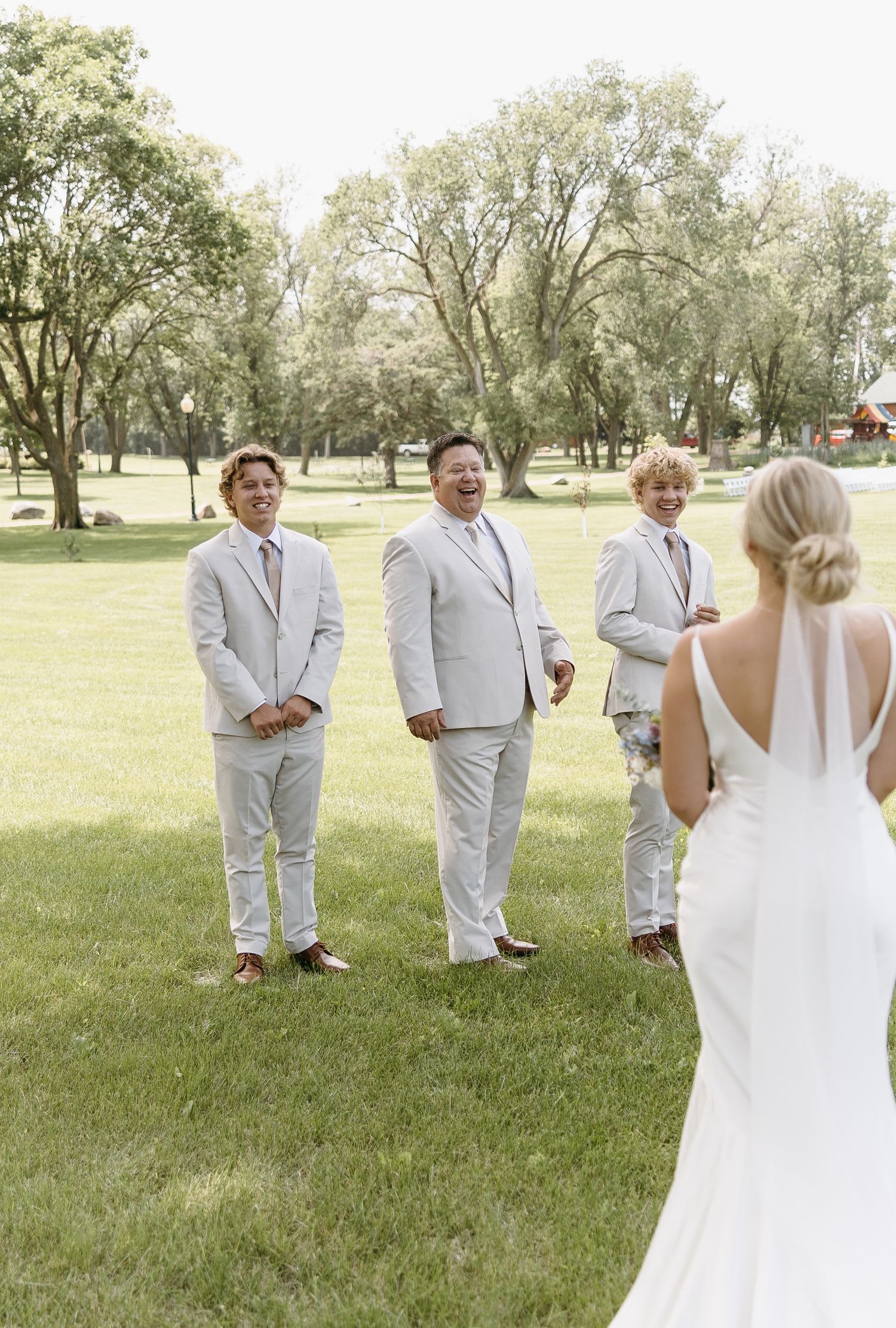 Wedding party members in light gray suits stand on grass greeting bride in elegant white dress.