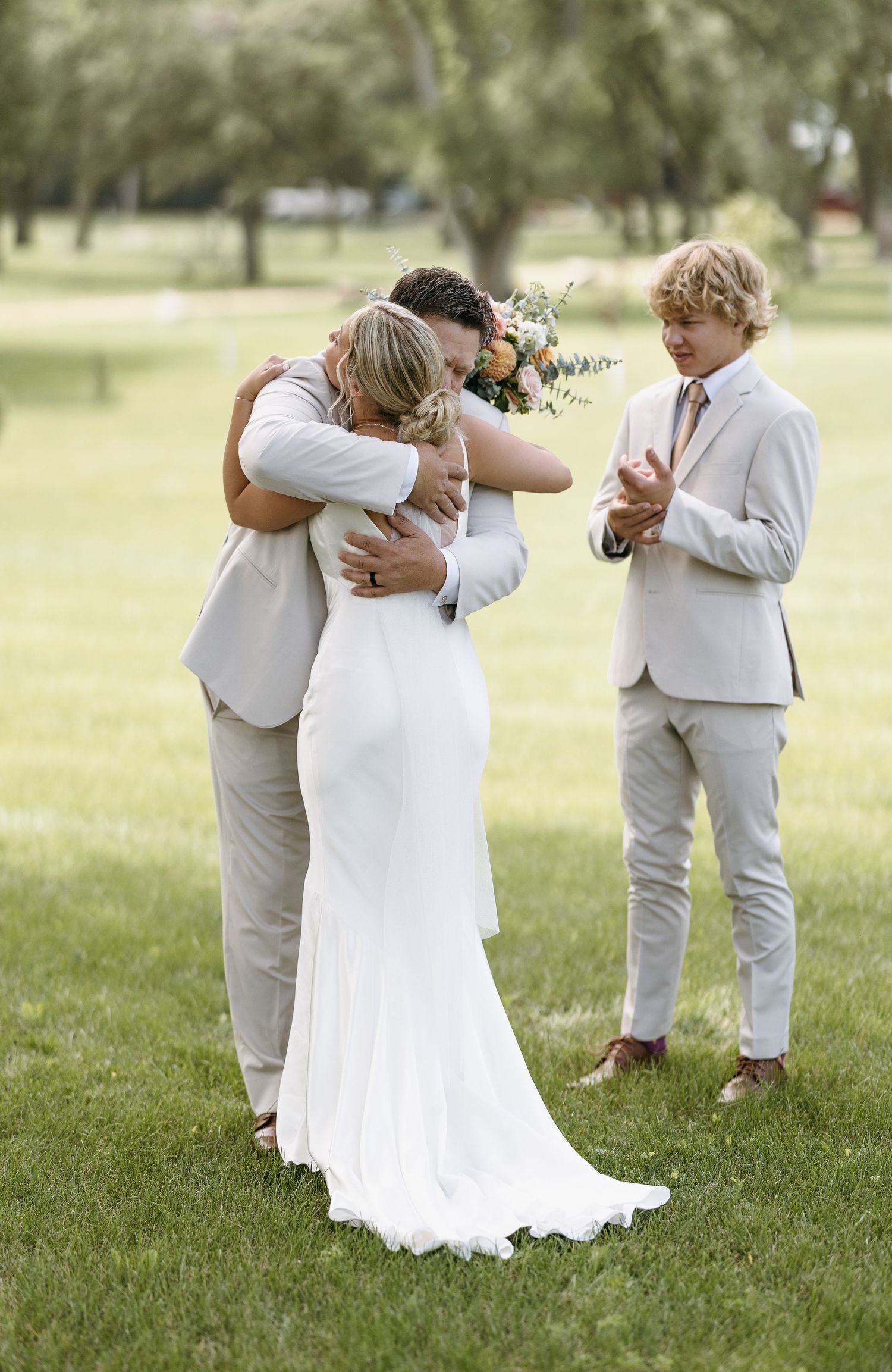 Emotional wedding moment captured outdoors with group embracing on green lawn.