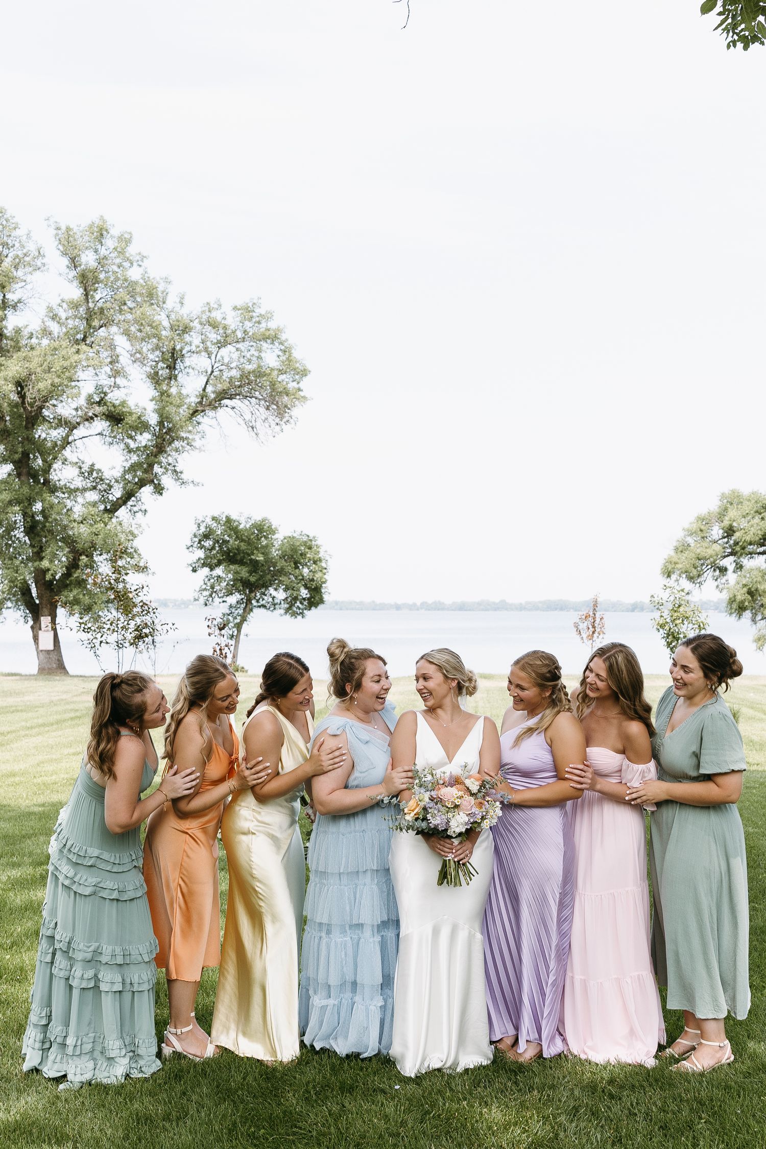 Bridal party in pastel dresses poses together by lakeside under trees.