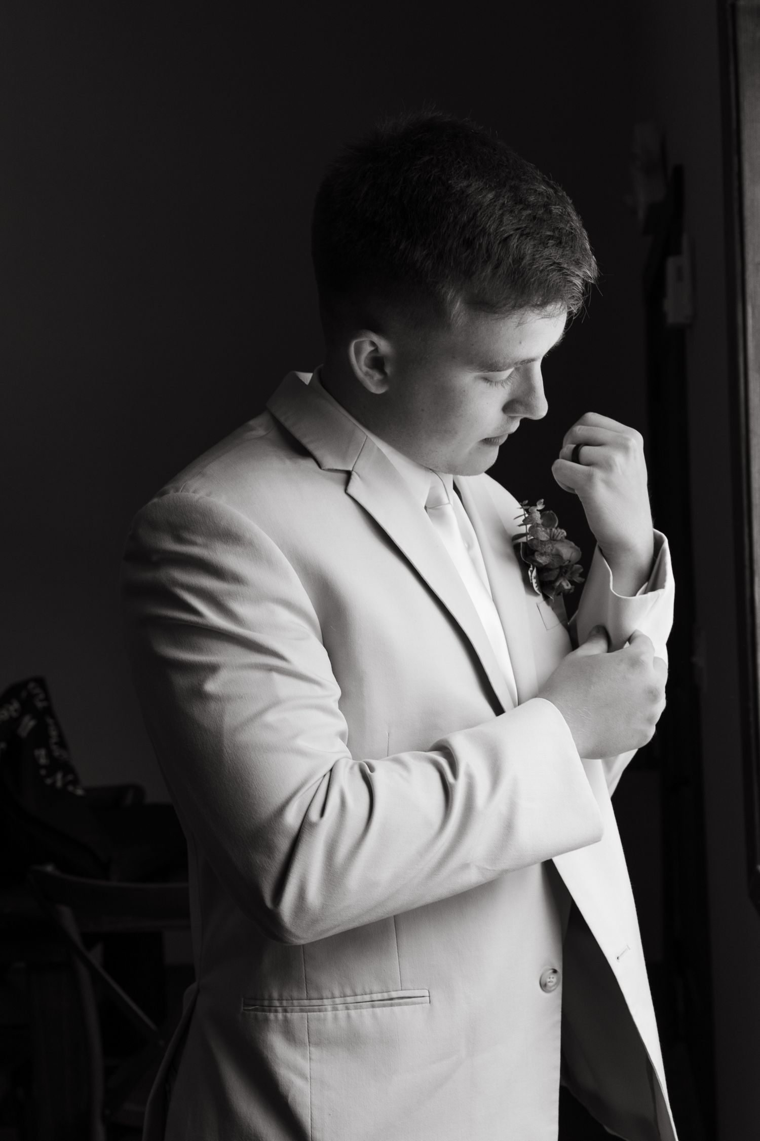 Black and white portrait of groomsman adjusting boutonniere in light suit.