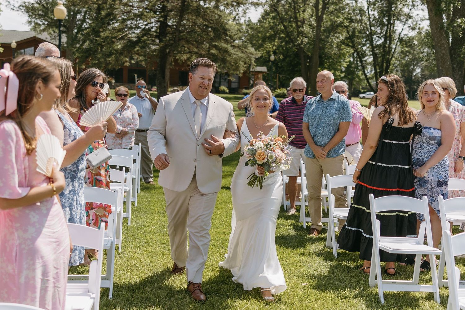 Wedding party walks down outdoor aisle with guests watching on white chairs surrounded by trees.