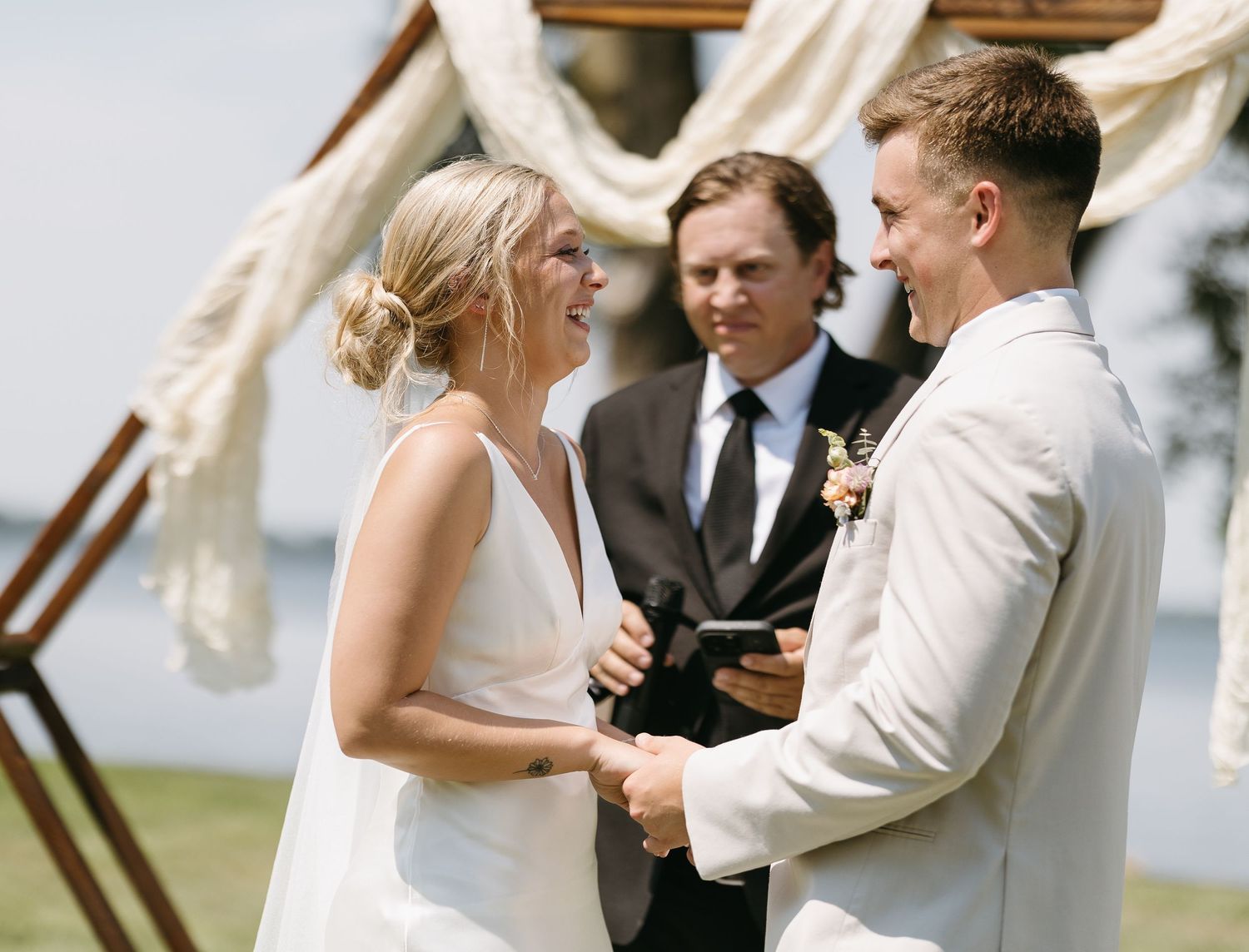 Outdoor wedding ceremony under wooden arch with white fabric draping and water view.