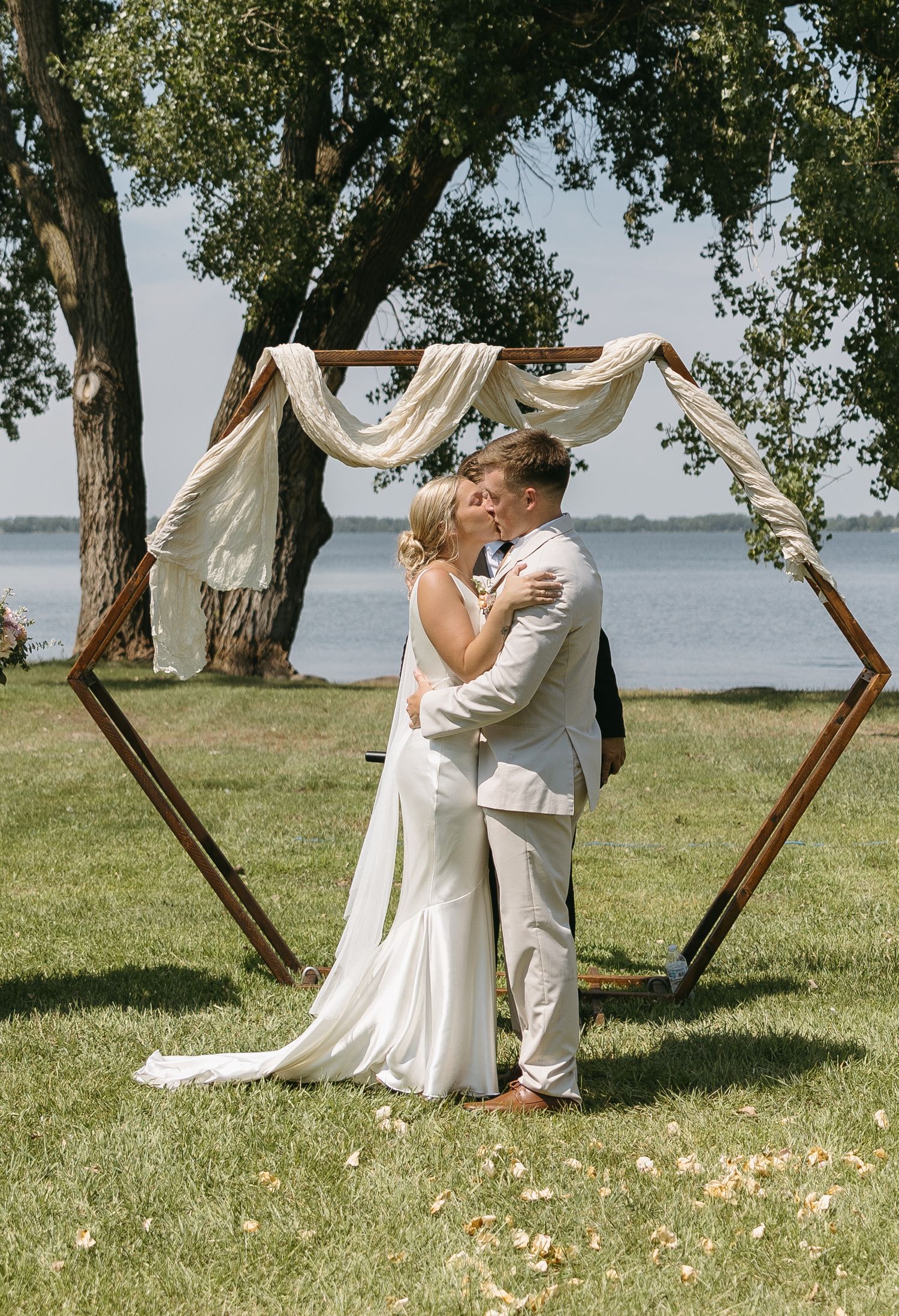 Couple embraces under hexagonal wooden arch draped with white fabric by lakeside.