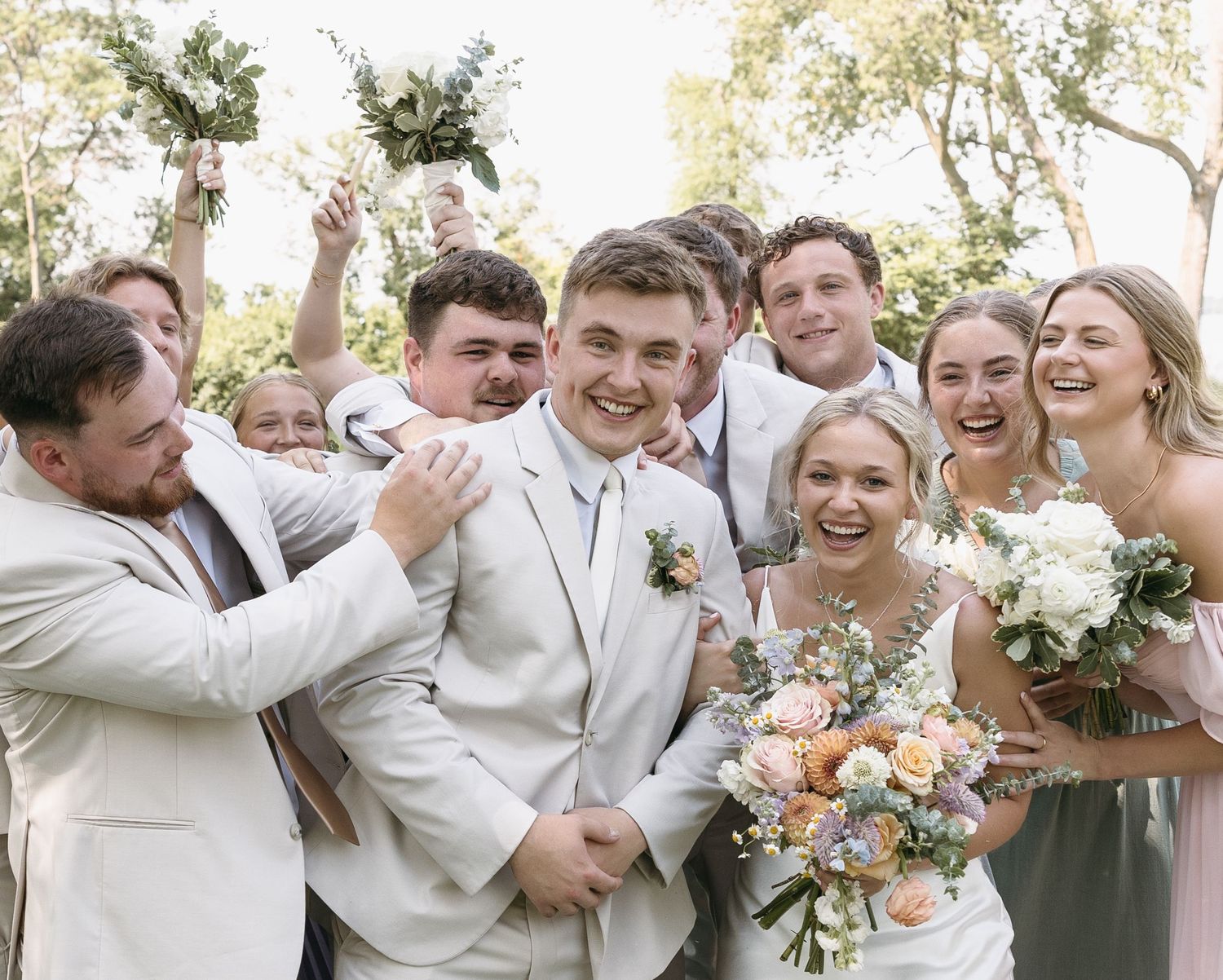 Wedding party shares laughter and celebration outdoors in light colored attire.