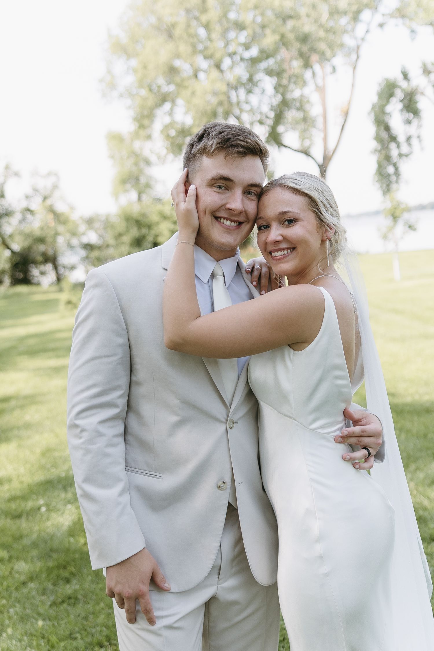 Couple poses outdoors in elegant white wedding attire against green landscape.