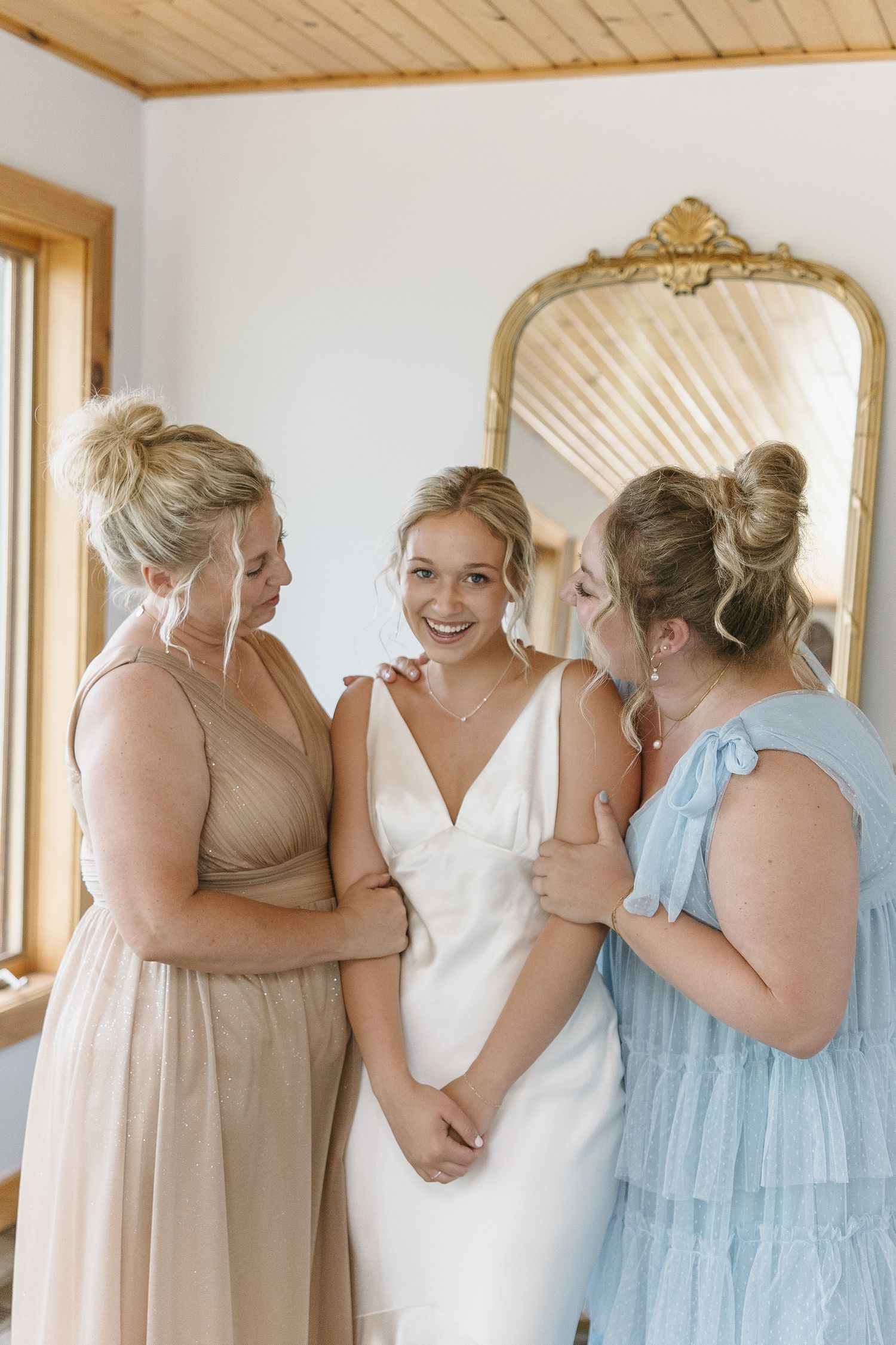Three women share a tender moment during wedding preparations in front of an ornate mirror.