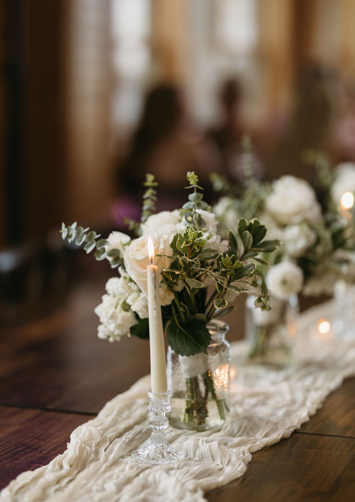Elegant wedding table centerpiece with white flowers, greenery and candles on lace runner.
