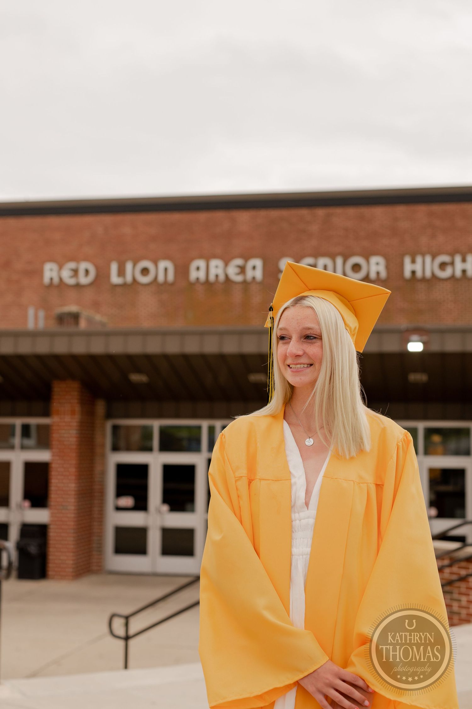 Graduate in yellow graduation attire smiles in front of high school entrance.