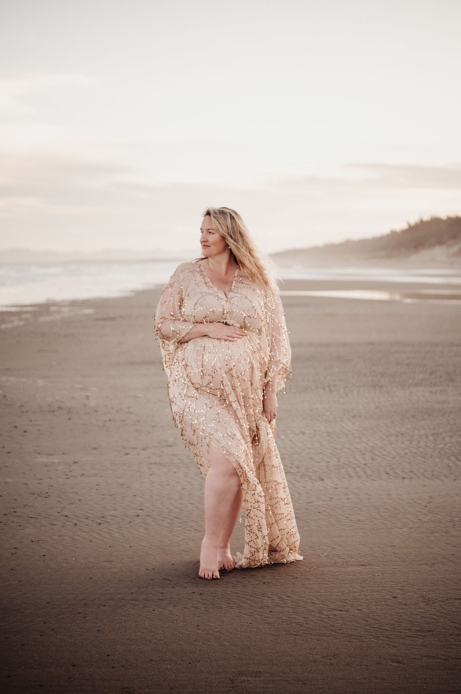 A figure in a delicate lace dress poses on a misty beach at sunset.