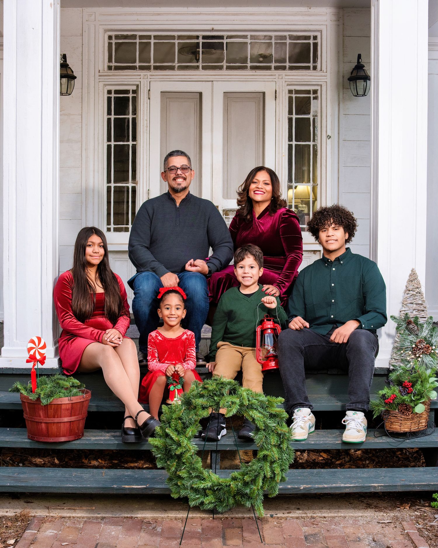 A family sits together on a front porch decorated with Christmas wreaths and decorations in a holiday themed photo.