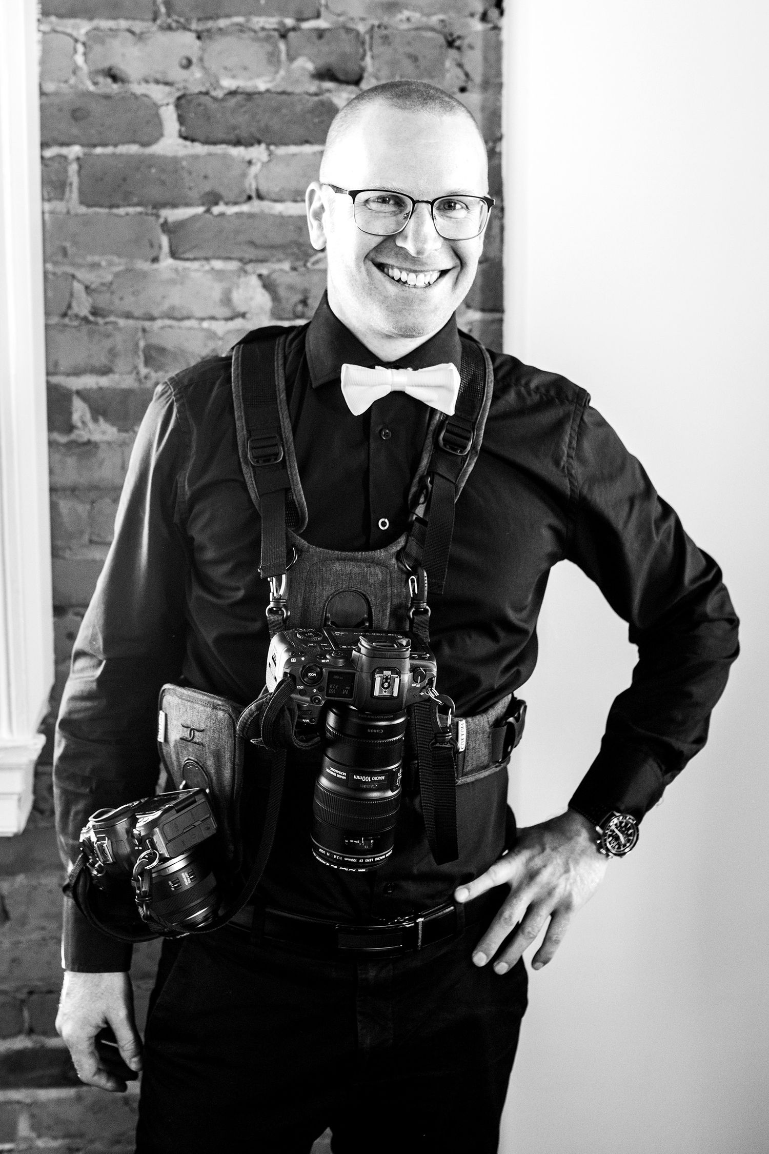 Portrait de Brophoto en tenue de soirée, équipé d'un système de harnais à double appareil photo, devant un mur de briques.