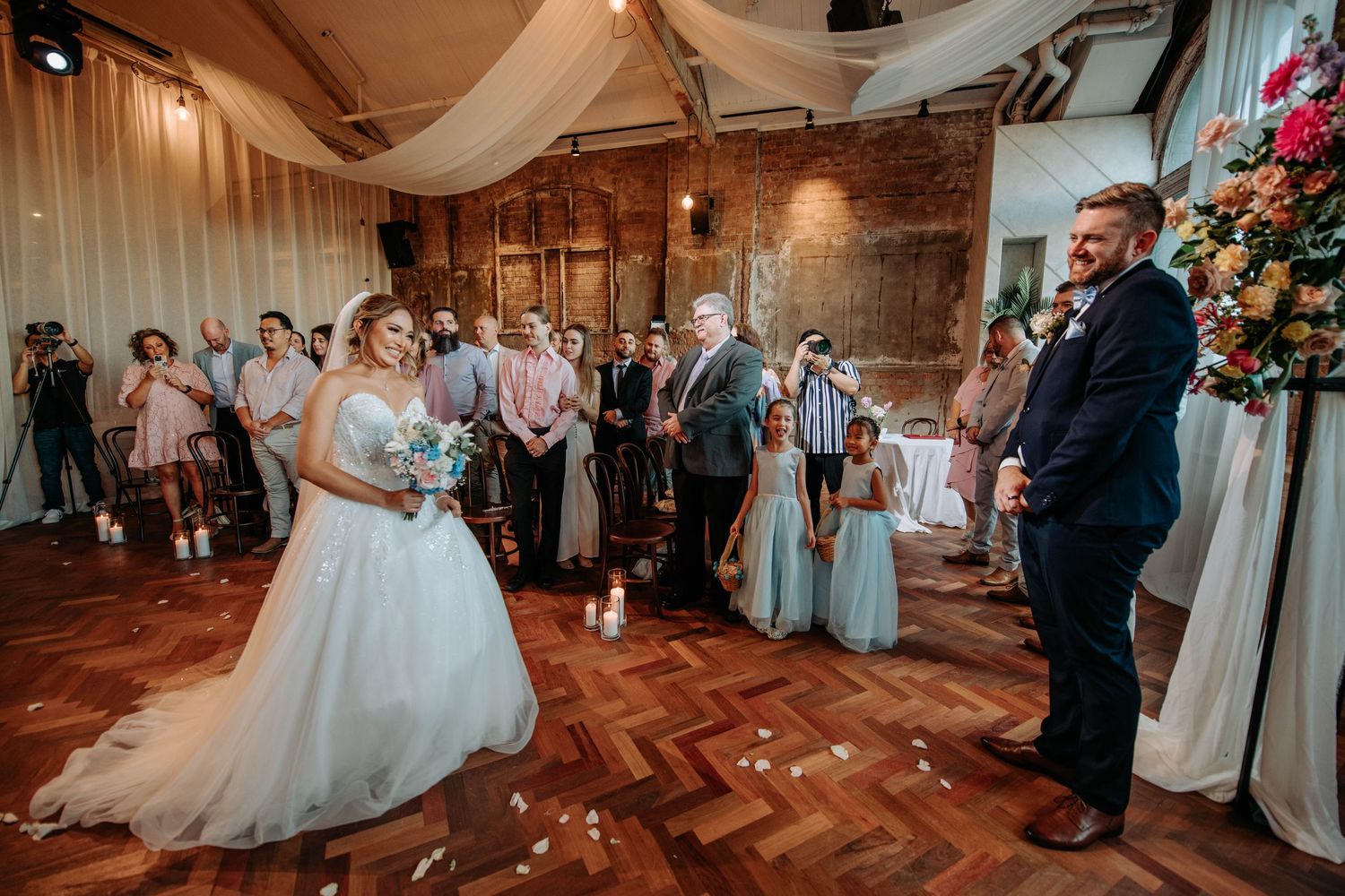 Newlyweds share first dance surrounded by guests in rustic venue with wooden floors.