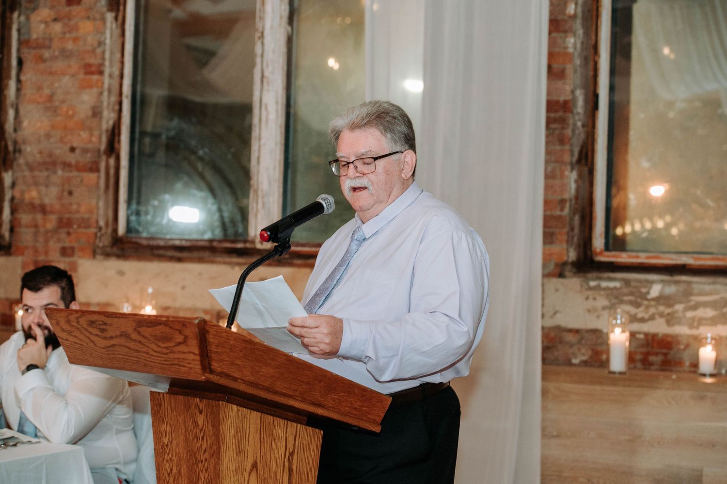 Speaker delivering a speech at a wedding reception podium with exposed brick walls.