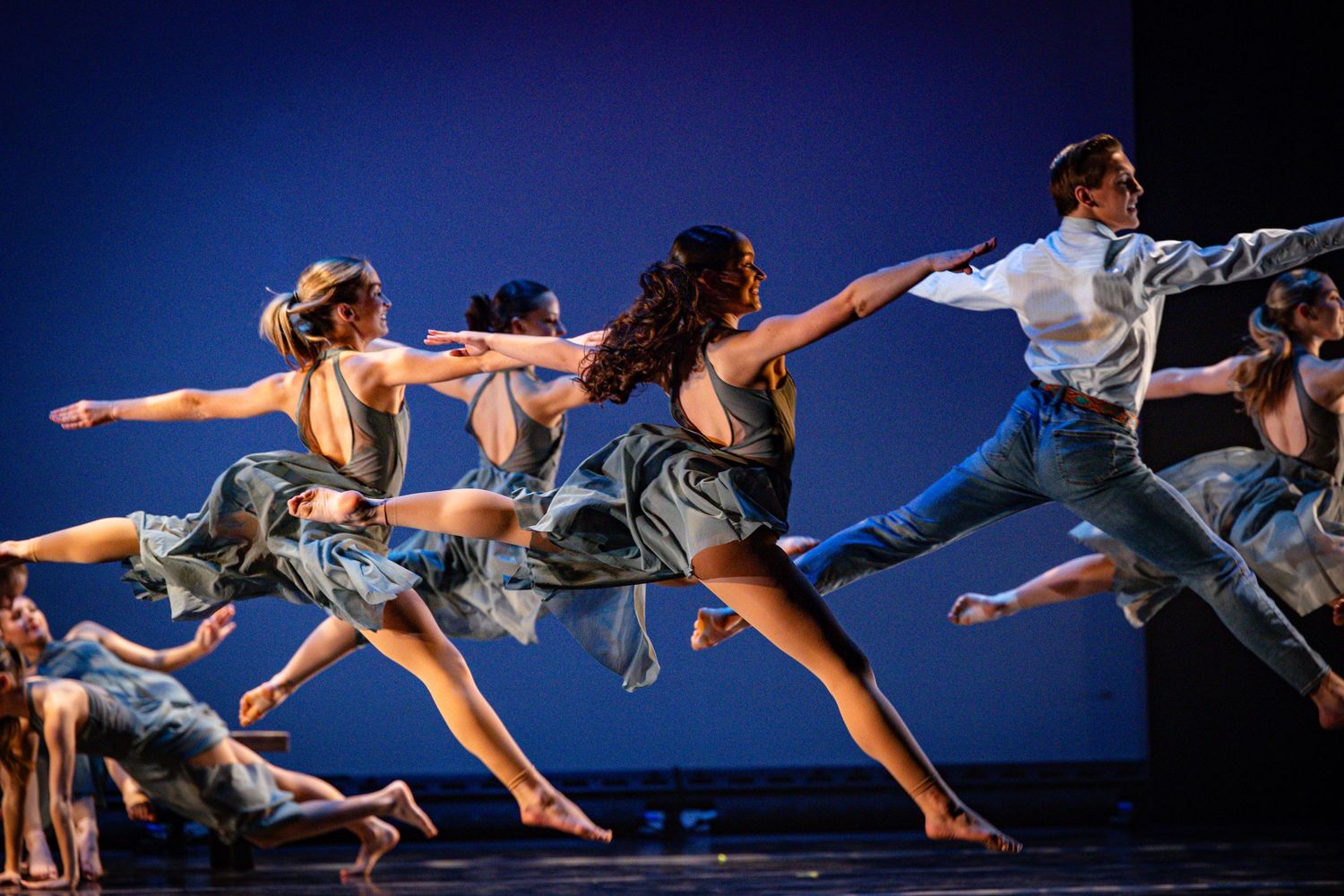 Modern dancers leap in synchronized formation against blue backdrop wearing casual costumes.