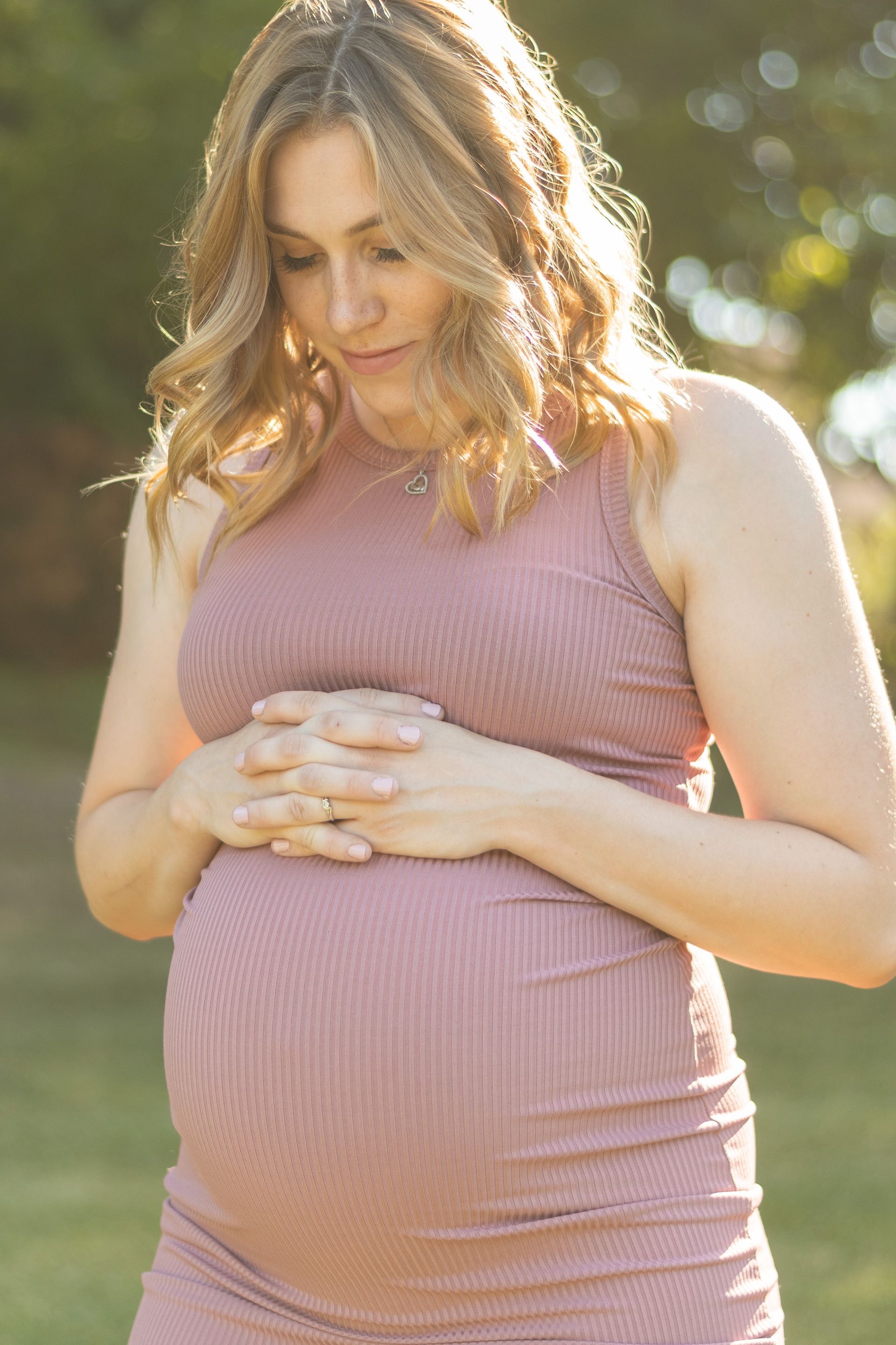 Pregnant woman in pink dress touches her belly during sunset maternity photoshoot.