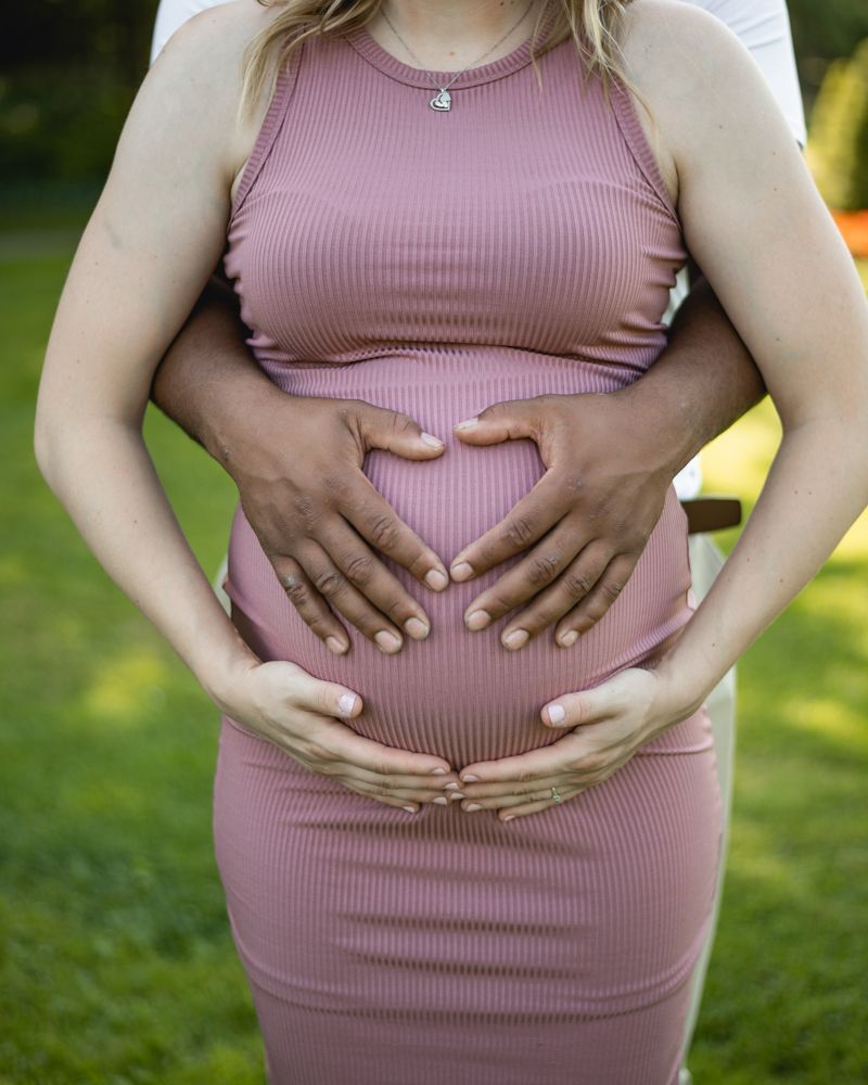 Close-up of hands forming heart shape on pregnant belly in pink dress.