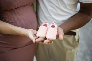 Couple holding tiny pink baby shoes against pregnant belly outdoors.