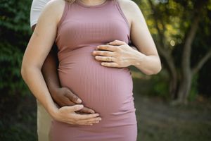 Loving hands embrace pregnant belly in form-fitting pink dress outdoors.