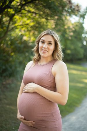 A woman in a dusty rose ribbed dress poses outdoors during a maternity photoshoot on a path with trees in background.