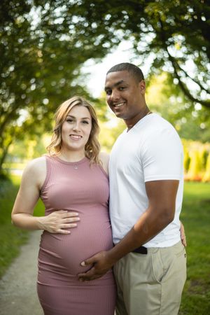 Expectant couple poses together outdoors on a path surrounded by greenery.