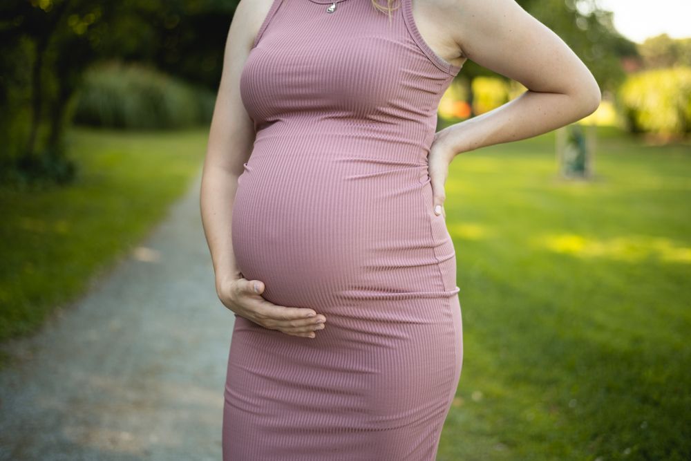 Side view of pregnant woman in pink dress cradling her baby bump outdoors.