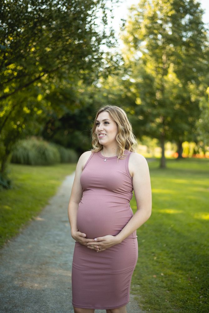 Maternity portrait of woman in pink dress standing on garden path with golden sunlight.