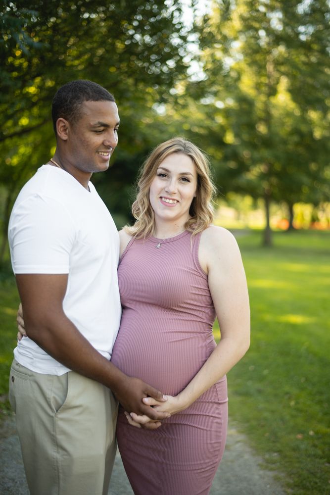 Expectant couple poses outdoors in a park setting with trees and sunlight in the background.