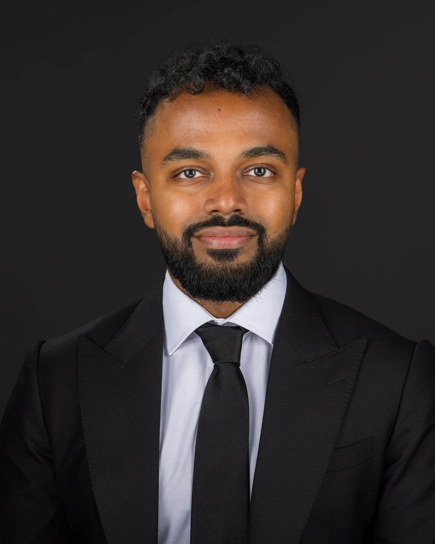 Professional headshot of someone wearing a black suit and tie against a dark background with soft lighting.