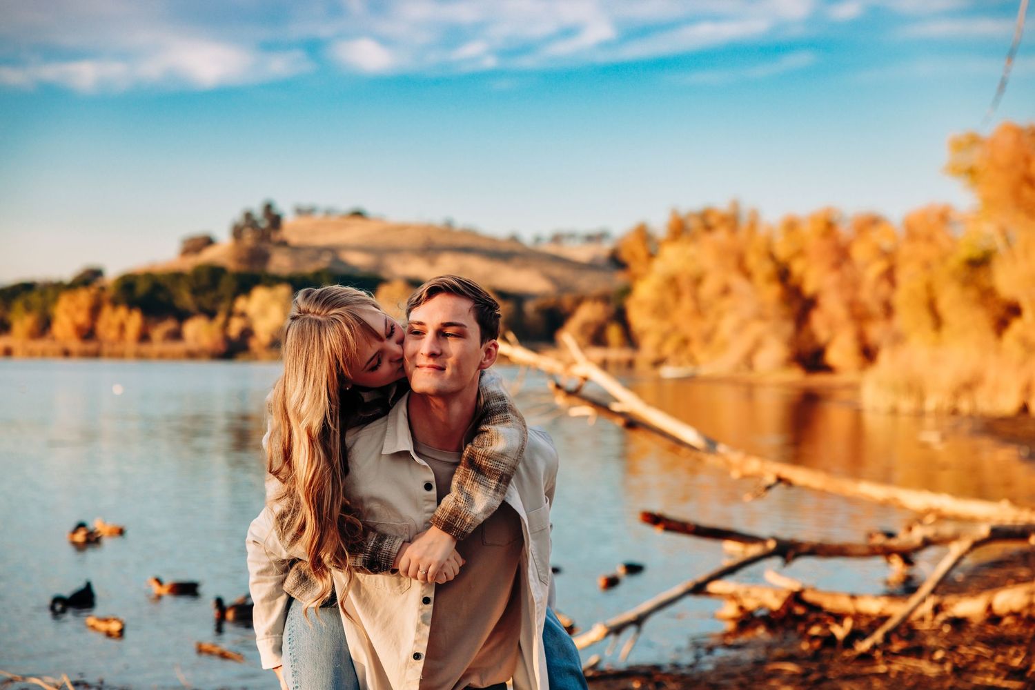 Playful couple embracing by lake with autumn trees in background.