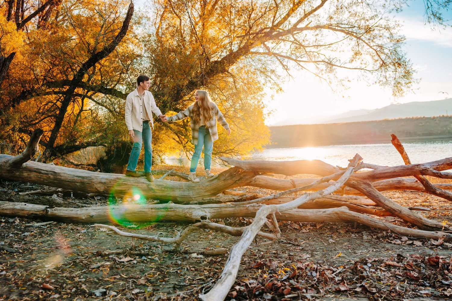 Couple walking on fallen logs by lake during golden hour sunset.