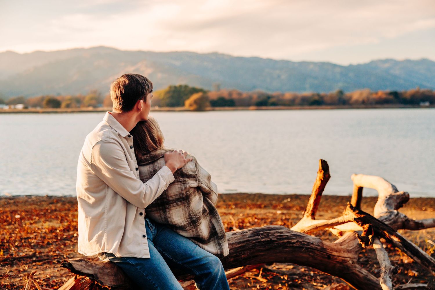 Couple enjoys autumn lakeside date with blanket and picnic setup against mountain backdrop.