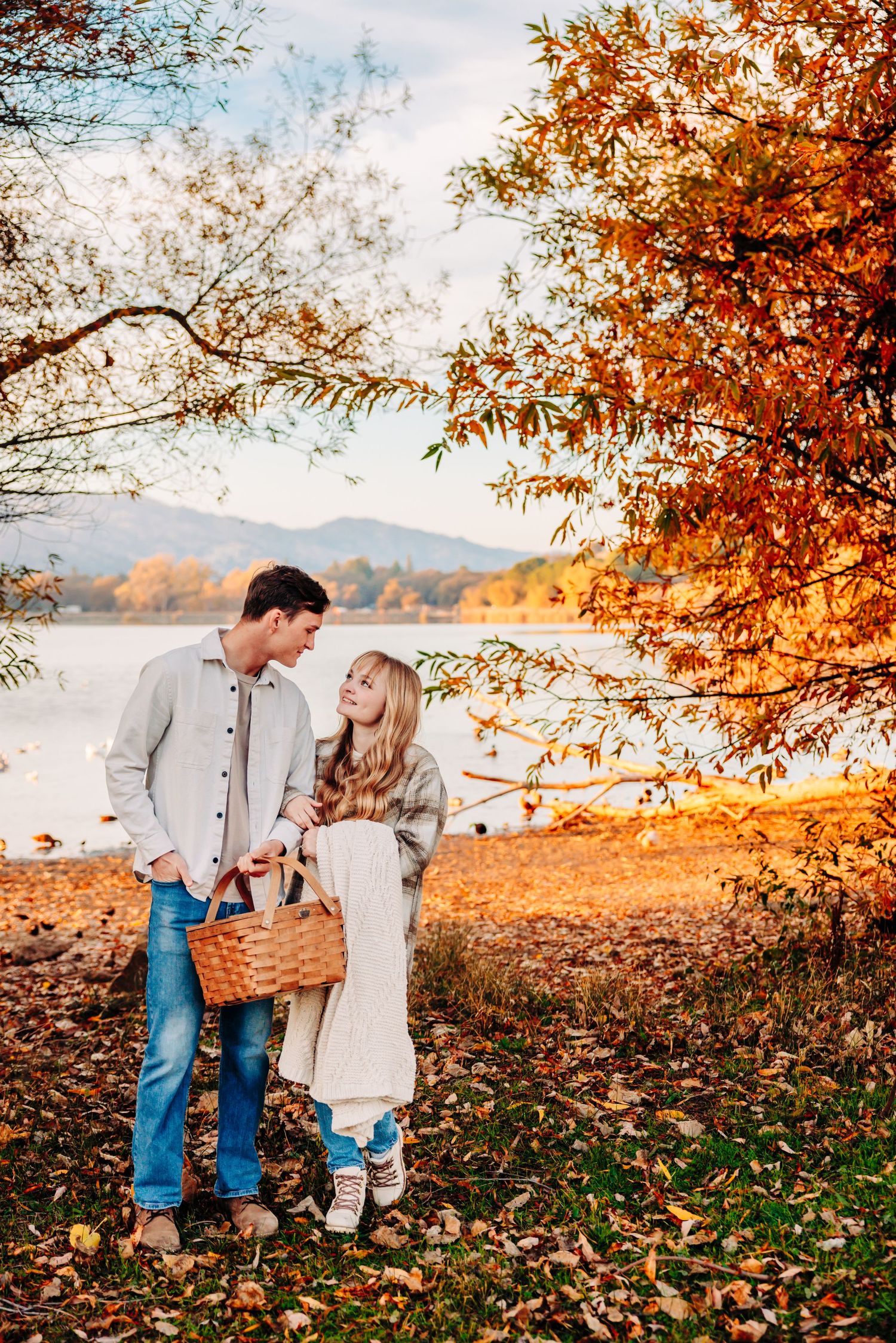 Couple holding picnic basket among fall foliage by serene lake.
