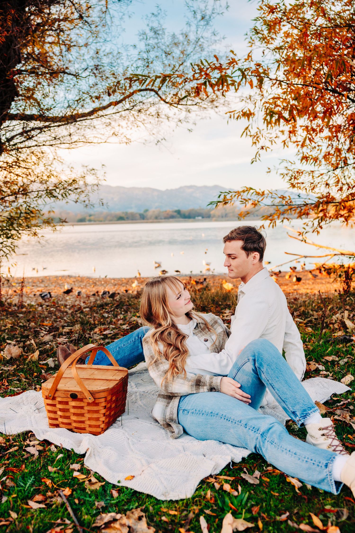 Picnic scene with couple and small dog on blanket under autumn trees by lake.