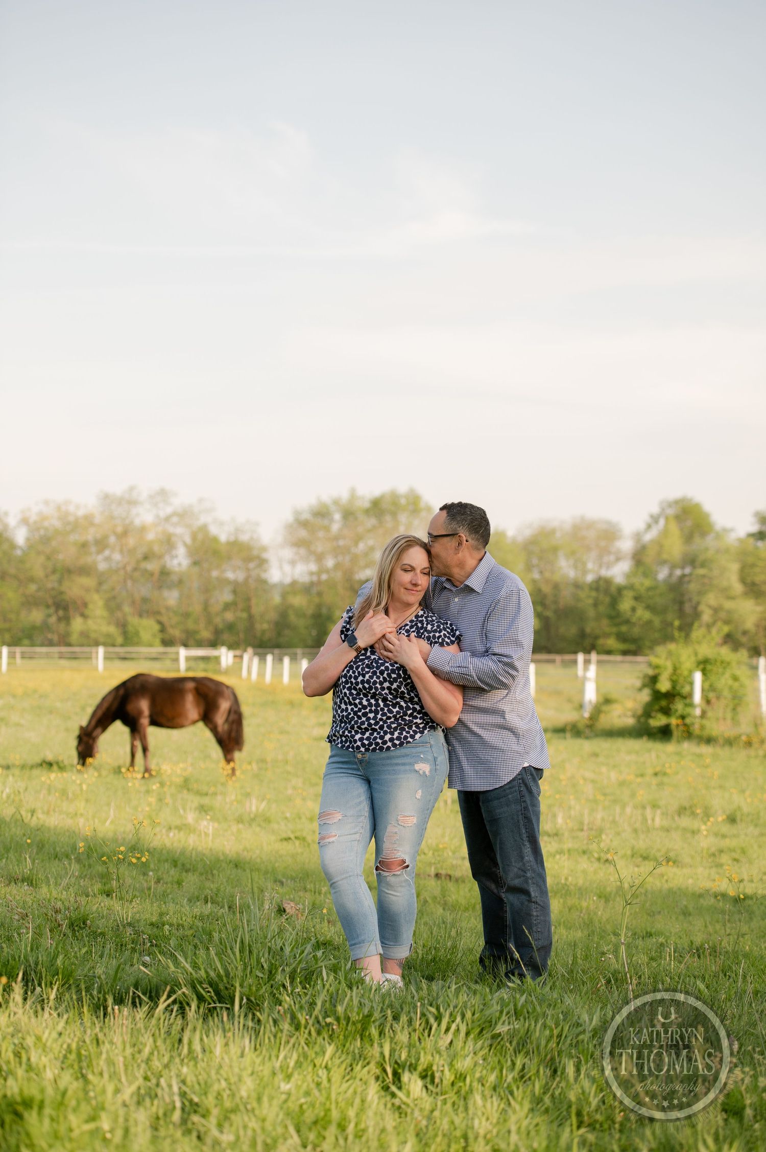 A tender moment between a couple in a pasture with their horse grazing in the distance.