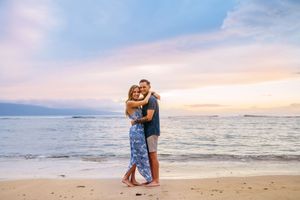 A couple stands together on a sandy beach with ocean waves and sunset in the background in lahaina maui for couples photography session