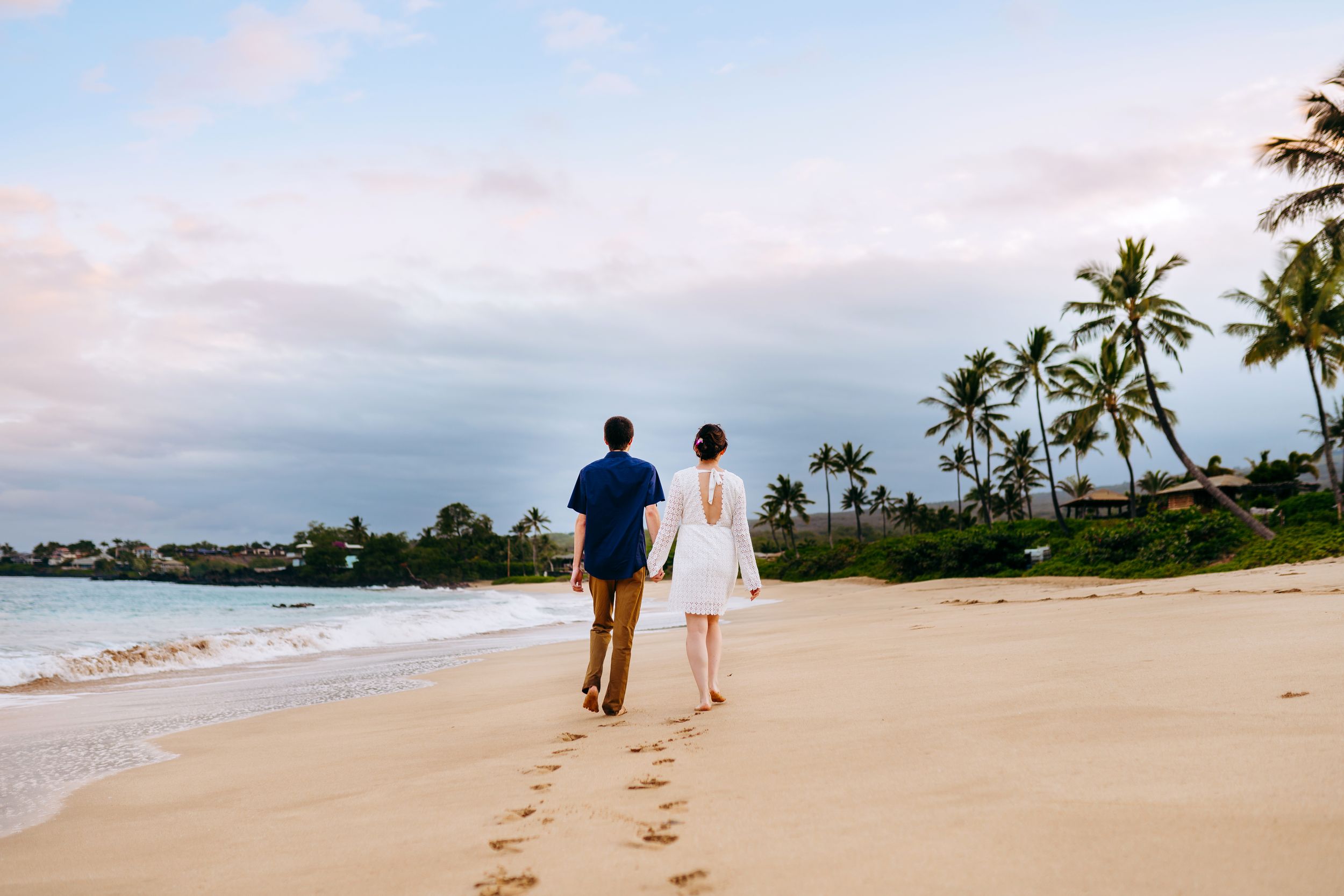 South Maui couples photography at sunset. Footprints trail behind a couple walking along a pristine beach with palm trees in the distance.