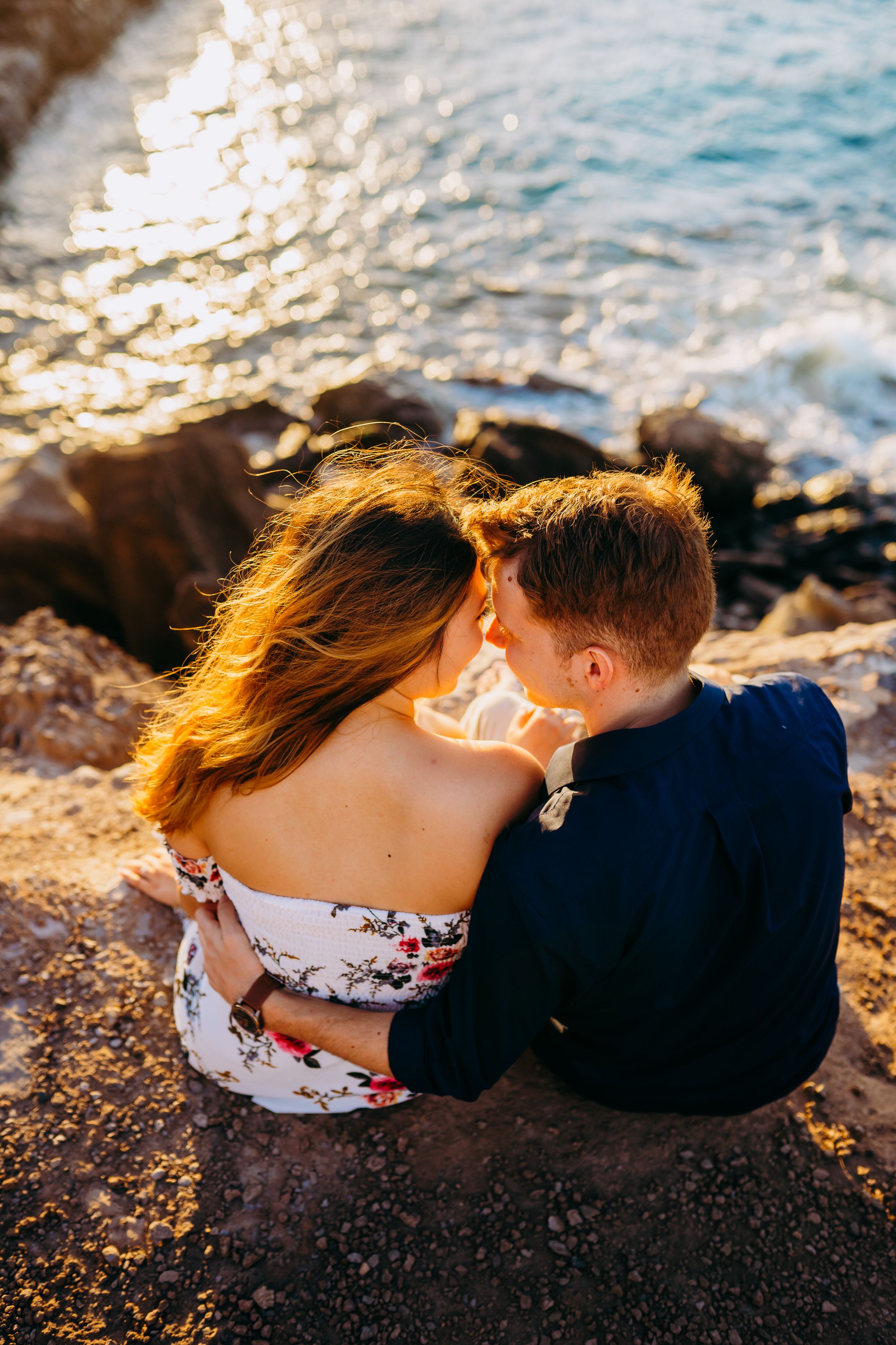 A romantic couple sits close together on a rocky beach at sunset with waves crashing behind them in kapalua maui for couples photography s
