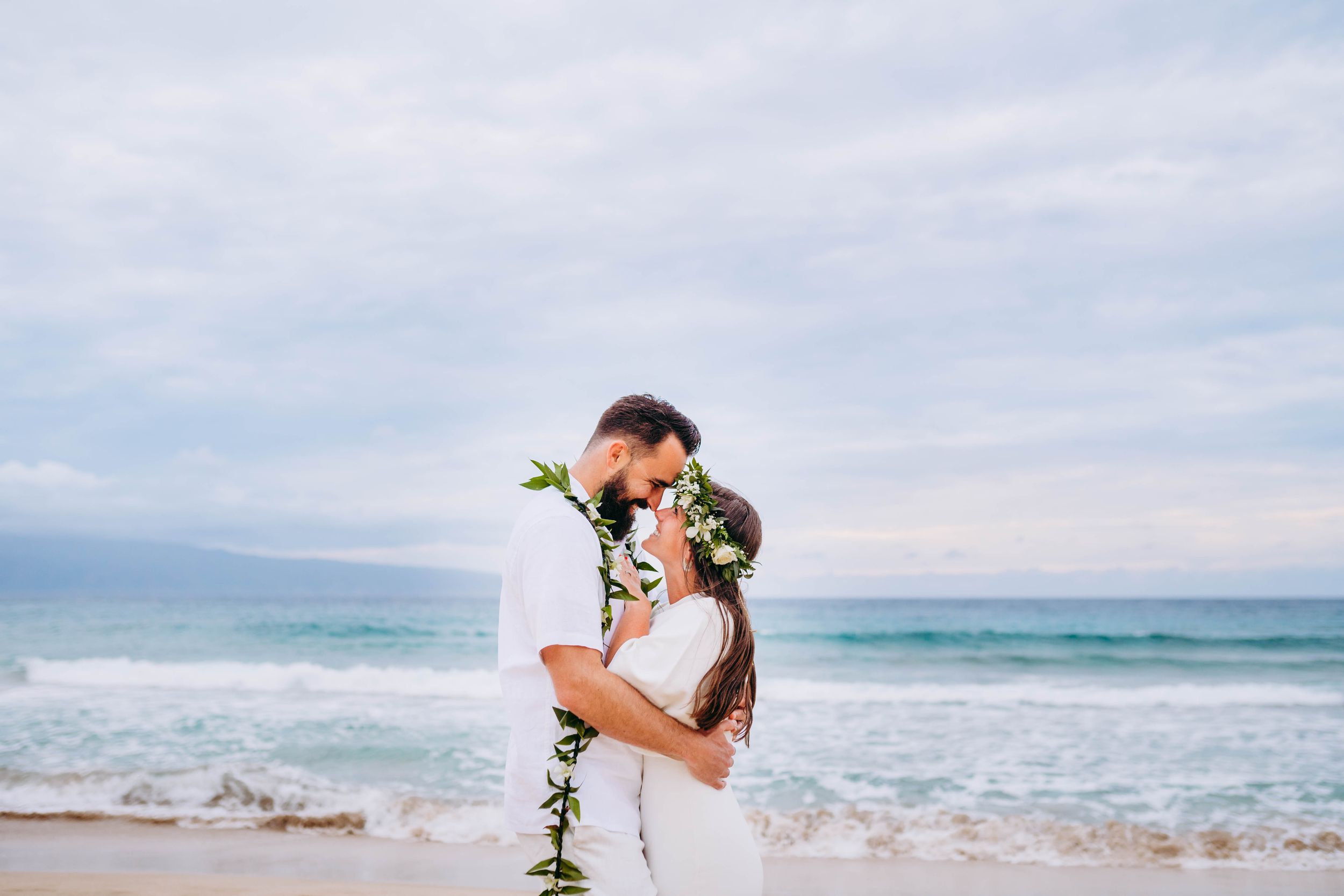 floral lei couples photography. Couple sharing an intimate moment on a tropical beach with waves and palm trees.