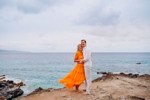 West Maui couples photography at sunset. A couple in flowing orange and white outfits walks along a rocky oceanside cliff.