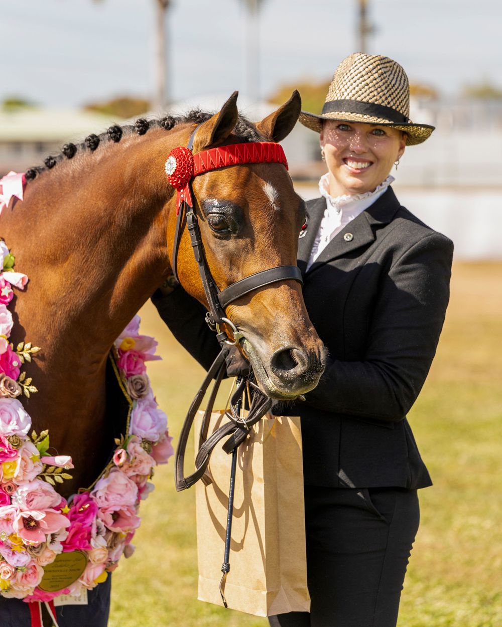 A bay horse wearing a red bridle and decorated with pink flowers poses with its handler in formal equestrian attire.