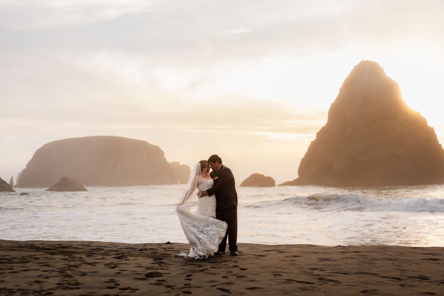 Bride and groom share a romantic kiss on a misty beach at sunset with dramatic sea stacks in the background.