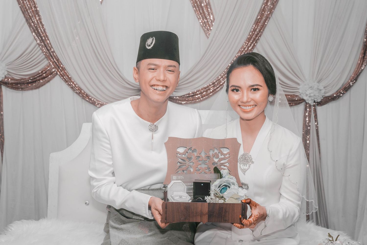 Wedding couple in white attire holding a decorative gift box against draped backdrop.