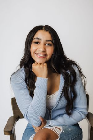 Portrait of young woman in blue cardigan and jeans sitting casually with hand under chin.
