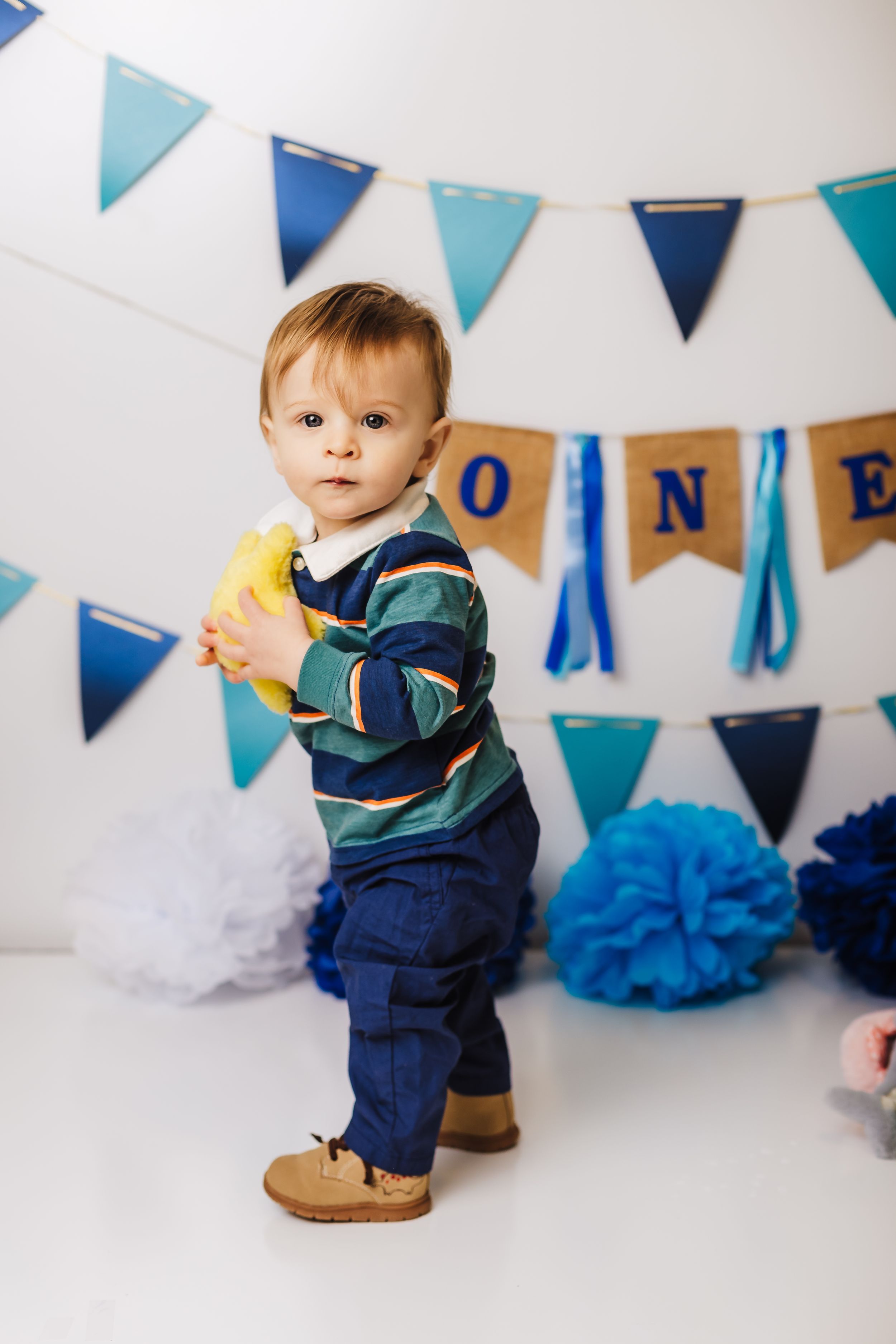 Toddler in striped shirt and jeans stands by blue birthday party decorations.