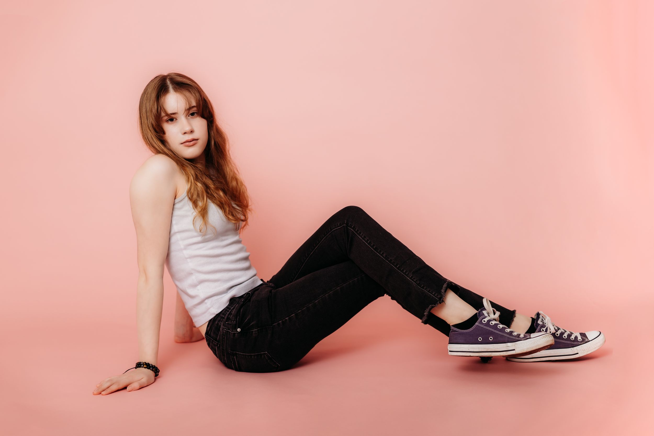 Young person in white tank top and black pants sitting on pink backdrop.