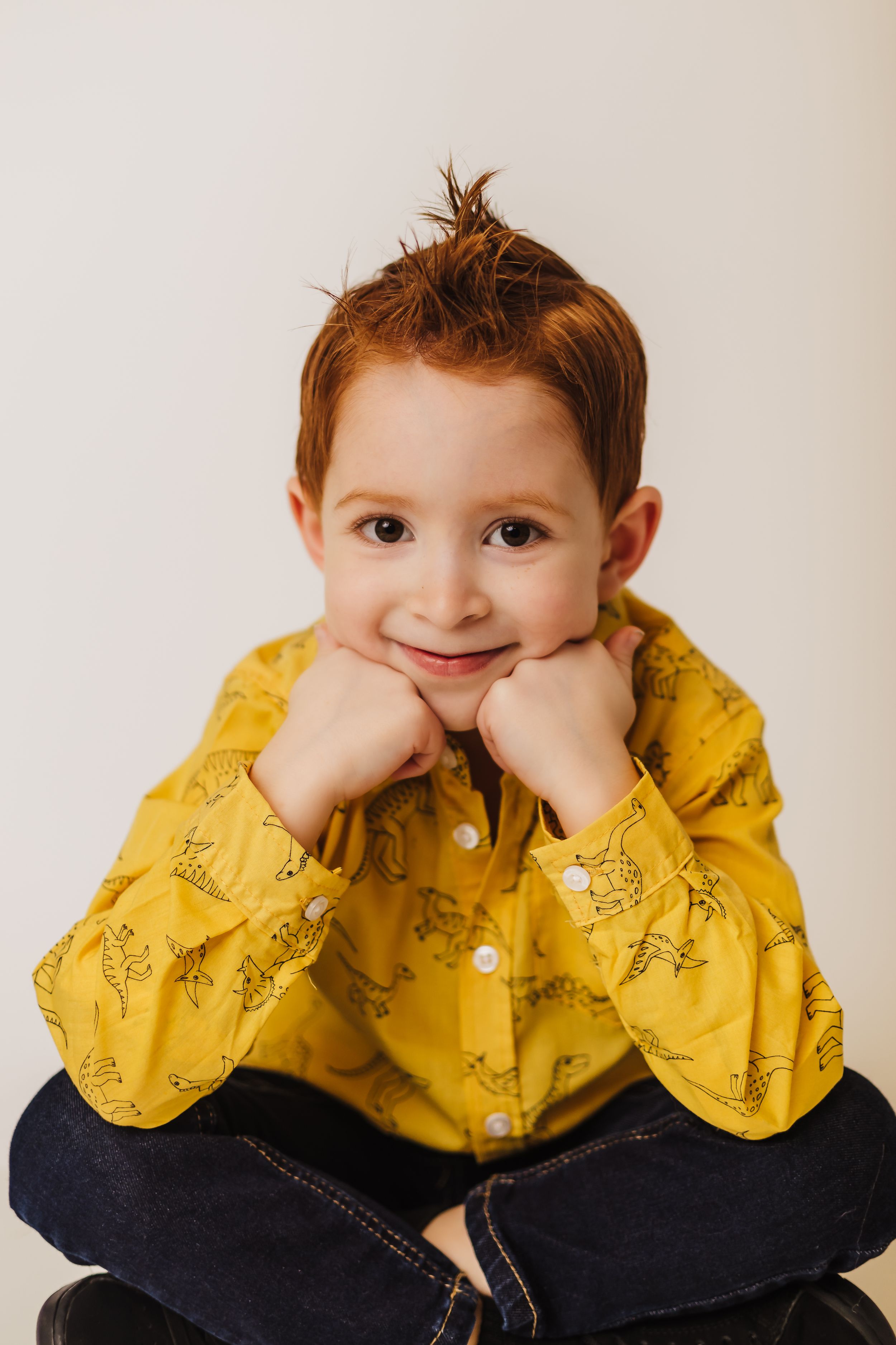 Child in yellow patterned shirt sits cross-legged with hands under chin against white background.