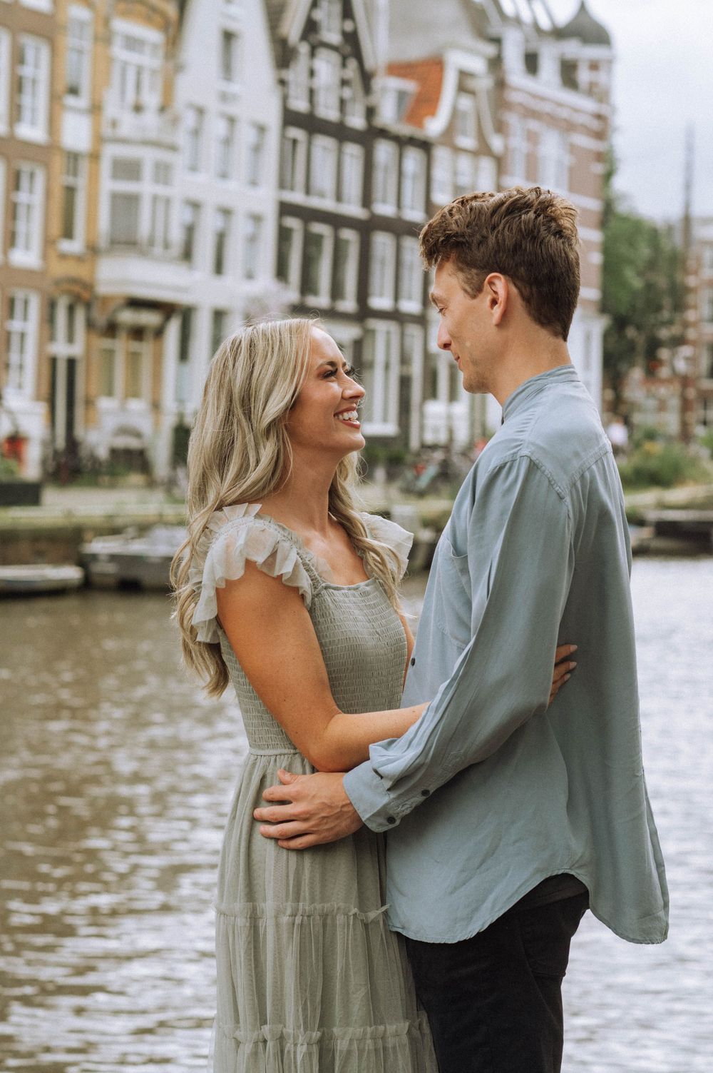 A couple shares a tender moment along an Amsterdam canal with historic buildings reflecting in the water.