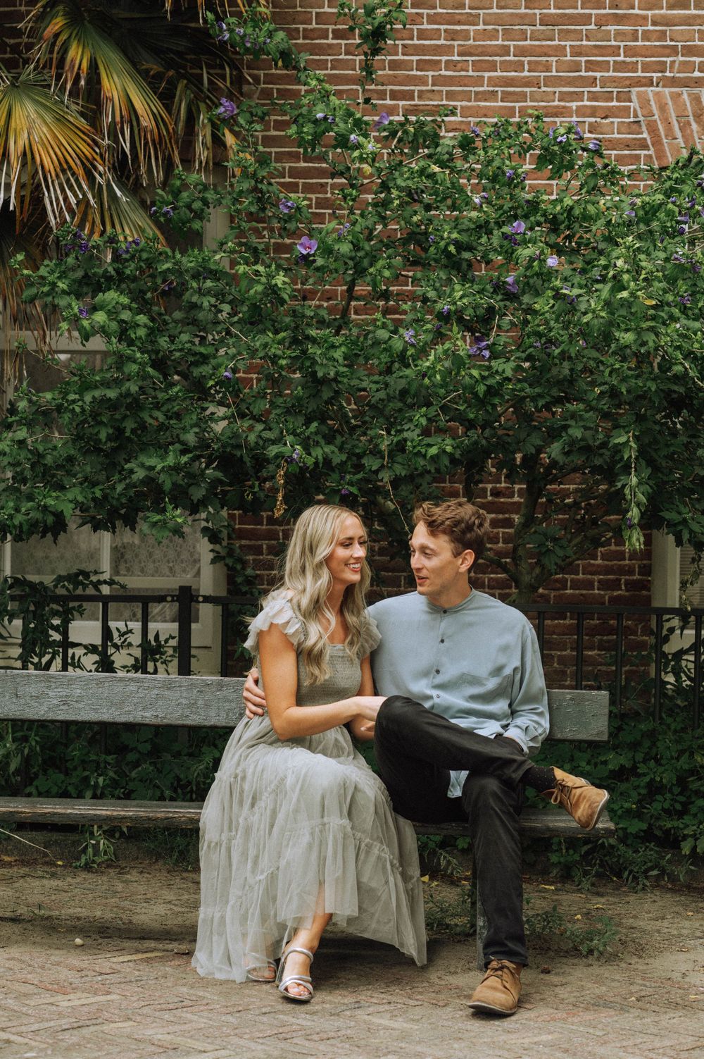 A couple sits together on a bench near a brick wall with lush green foliage.